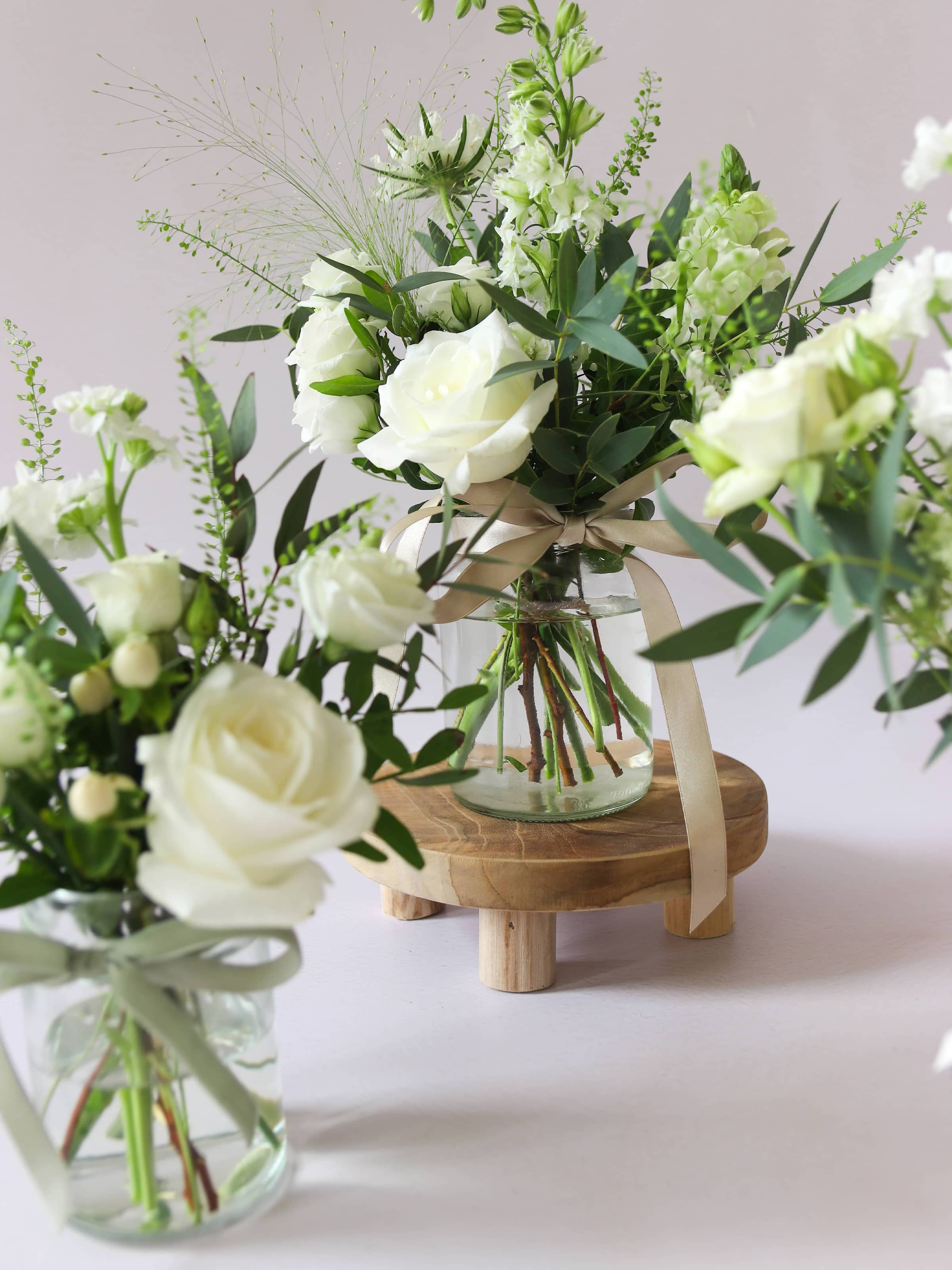 Two jam jars with white and green flowers sitting on a wooden stool against a light background